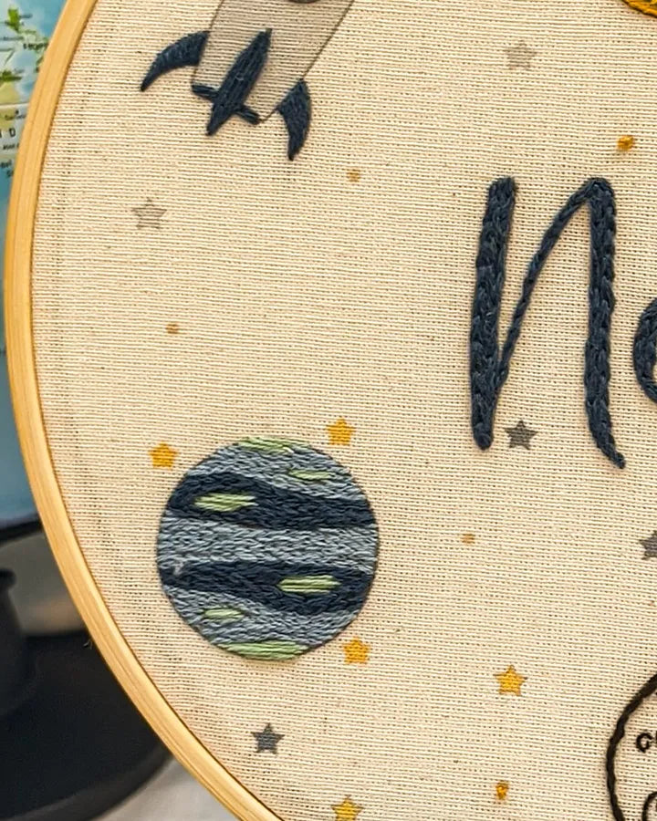 Embroidery hoop with embroidered design of a planet, stars, and text.
