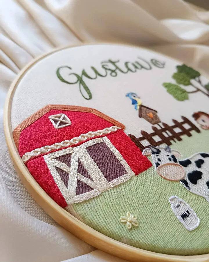 Embroidery hoop with farm scene including a red barn, cow, and other elements on a beige background.