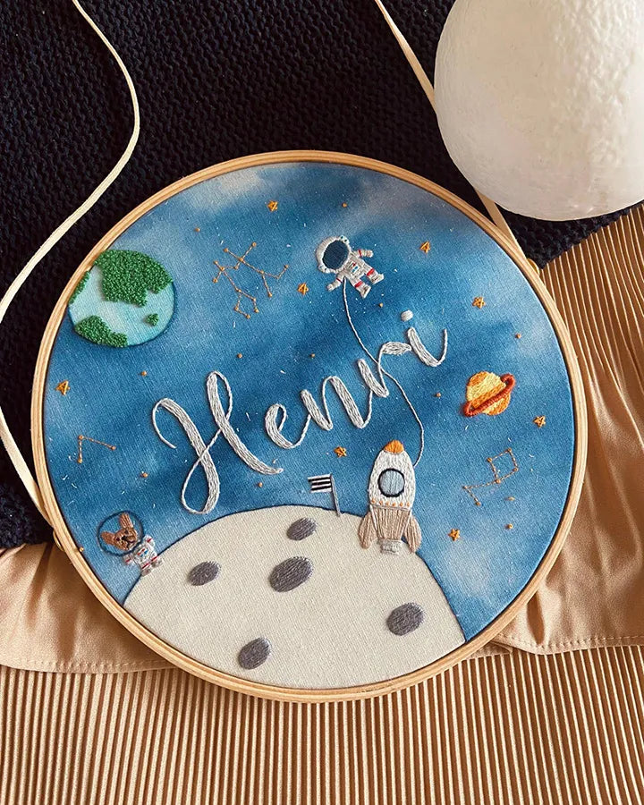 Embroidery hoop with space-themed design featuring a rocket, moon, and Earth.